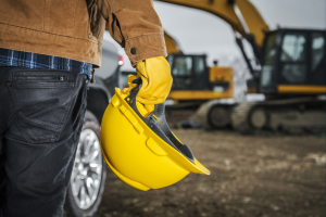 Person wearing work gloves and holding a yellow hard hat with construction equipment in the background